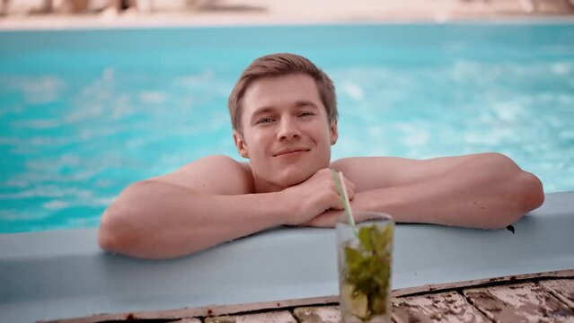 Shot of handsome happy man folded arms at swimming pool corner outdoors. Smiling boyfriend recreation in blue water enjoying his cold cocktail mojito. Satisfied guy on resort