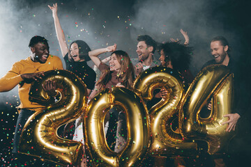 Group of joyful young people celebrating 2024 New Year while throwing confetti in night club