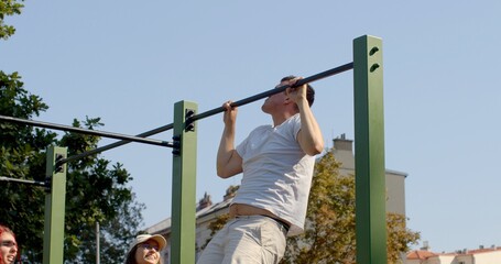 Fototapeta premium Young man engaging in physical activity by performing pull-ups on horizontal bar accompanied by friends, scene reflects urban lifestyle of active young people. Physical fitness and exercise outdoor.