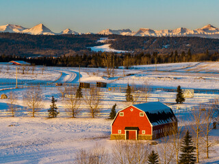 Red barn in a snow-covered field glowing with the warm light of sunrise with foothills and snow-covered mountains in the background and blue sky, West of Calgary, Alberta; Alberta, Canada