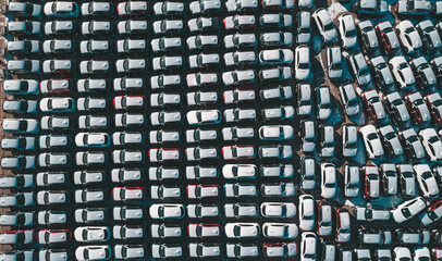 Fototapeta premium Aerial top down view of new cars parked in car parking lot. Car dealer parking lot full of new automobiles. New cars lined up for import and export business.