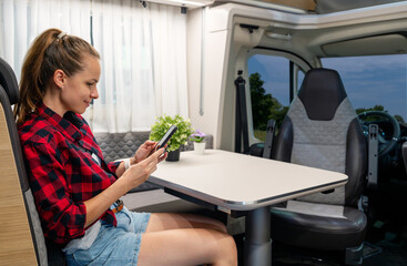 Young woman sits at the table inside of her camper van and using her mobile phone during night parking.