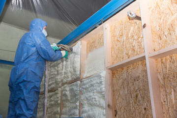 The builder in a blue suit applies a construction foam from the gun to the wall of a wooden house