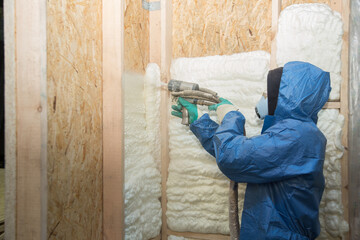 The builder in a blue suit applies a construction foam from the gun to the wall of a wooden house