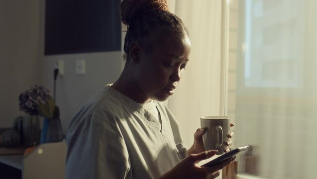 Medium tracking shot of African American doctor in medical uniform walking around apartment, drinking coffee, browsing on smartphone, then checking time, taking keys and ID badge, leaving for shift