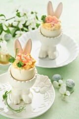 Stuffed or deviled eggs with yolk, shrimp, pea microgreens with paprika in rabbit-shaped stand for easter table decorate fresh cherry or apple blossoms on light background. Traditional dish for Easter