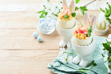 Stuffed or deviled eggs with yolk, shrimp, pea microgreens with paprika in rabbit-shaped stand for easter table decorate fresh cherry or apple blossoms on light background. Traditional dish for Easter