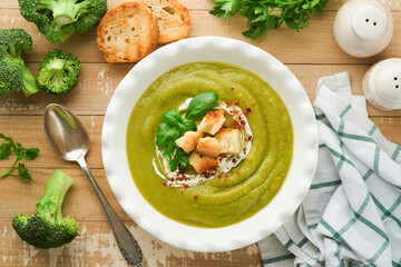 Broccoli cream soup. Fresh homemade broccoli soup in white plate with seasonings, parsley, basil and pieces of toasted bread on rustic white wooden background. Healthy, diet or baby food. Top view.