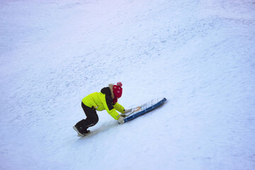 A persistent little boy pushes a sled up a hill so that he can then slide down on a sled.