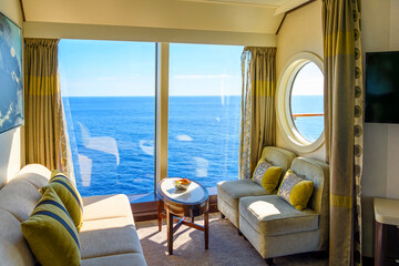 A bowl of fruit sits on a table in a luxury cruise ship cabin with a large window and porthole to balcony as it sails on a blue Mediterranean Sea.