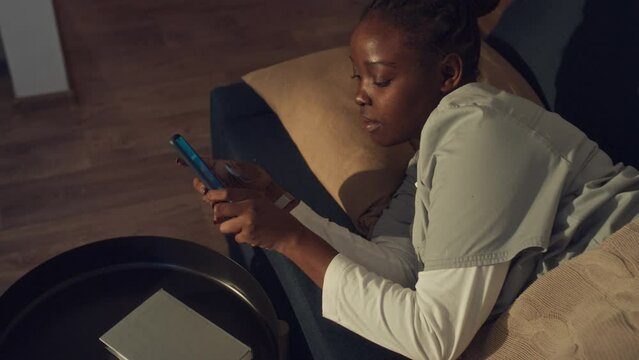 Medium top shot of black female hospital worker in uniform sleeping on couch at home, alarm clock beeping on smartphone, woman waking and getting up to go to shift