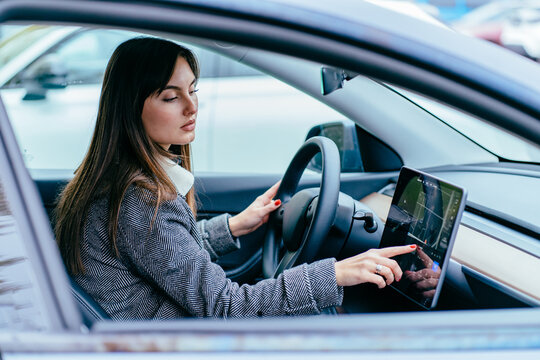 Young woman sits behind wheel in modern electric car wheel in car and uses an electronic dashboard, tablet computer. Girl is traveler looking for a way through navigation system.