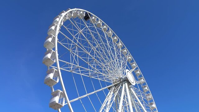 Large white spinning ferris wheel against at clean blue sky background 4K. Sunny day. Outdoor, event, park, fun fair, city, cab, object, rotate, ride, side, scene, view, motion, ultra hd. ProRes 422HQ