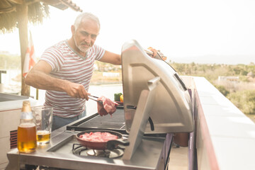 Mature man grilling steaks on a gas grill on his penthouse terrace - obrazy, fototapety, plakaty