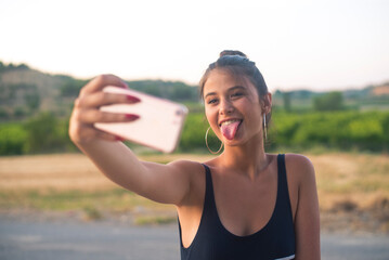 Portrait of teenage girl taking selfie with smartphone while sticking out tongue - obrazy, fototapety, plakaty