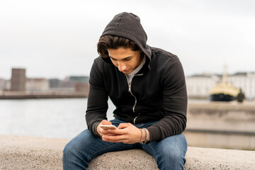 Denmark, Copenhagen, young man sitting on wall at the waterfront using cell phone