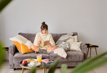 Woman reading book while sitting on sofa against wall at home during winter
