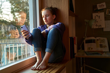 Girl sitting barefoot on window sill in the evening looking at smartphone