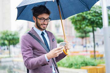 Fashionable businessman with umbrella checking his cell phone in the city
