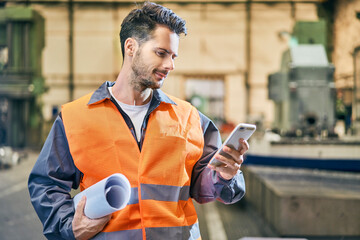 Smiling man holding blueprints and looking at cell phone in factory