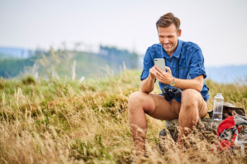 Smiling man resting and checking his cell phone during hiking trip