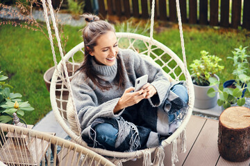 Happy young woman using smart phone while sitting on swing in garden
