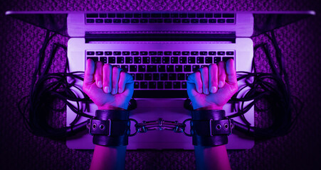 female sexy hands in bdsm handcuffs with laptop in neon light on dark background