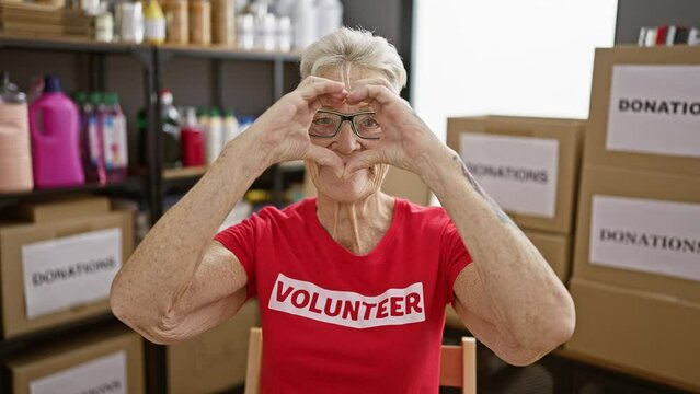 Confident grey-haired senior woman volunteer joyfully making a heart gesture in charity center, displaying love in service