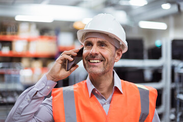 Smiling man in factory hall wearing safety vest and hard hat talking on cell phone