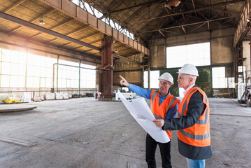Two men with plan wearing safety vests talking in old industrial hall