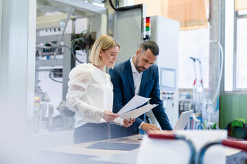 Businessman and young woman with papers and laptop talking in a factory