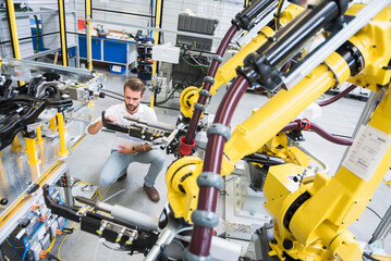 Full length of young engineer crouching while examining computer-aided machinery in automated industry - obrazy, fototapety, plakaty