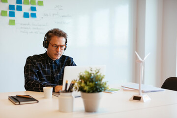 Businessman wearing headphones using laptop at desk in office - obrazy, fototapety, plakaty