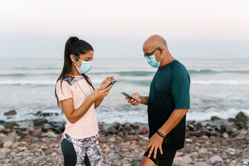 Woman and senior man with face mask using phone while standing against sea