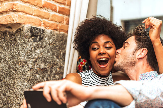 Close-up of man kissing cheerful girlfriend while taking selfie with her in coffee shop