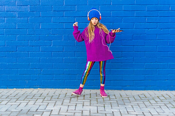 Girl wearing blue cap and oversized pink pullover listening music with headphones in front of blue wall