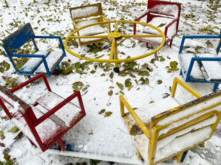 A round children's carousel with six seats in yellow, blue and red against a background of snow strewn with fallen dry leaves. Windy weather. Playground. Autumn season.