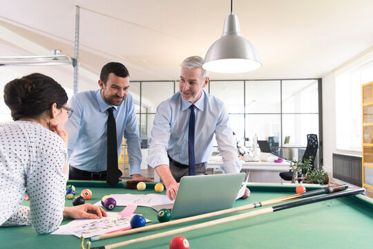 Business people standing at pool table with laptop, discussing investment strategy