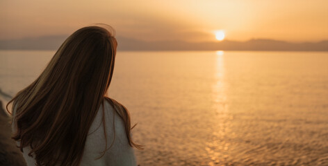 Scenic back view of a young woman with long, flowing hair, immersed in the breathtaking beauty of the sea and the setting sun