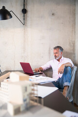 Businessman in a loft using laptop with documents and architectural model on table