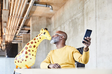 Mature businessman sitting at desk in office with cell phone kissing giraffe figurine