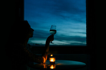 Silhouette birthday woman sitting with wineglass by window at home during sunset