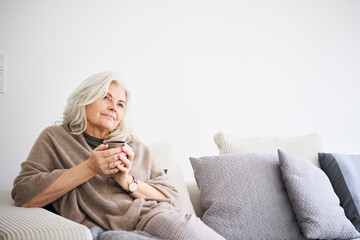 Thoughtful retired woman holding coffee cup while sitting on sofa against white wall at apartment