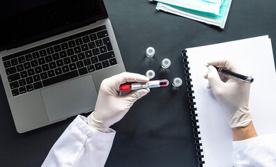 Doctor with COVID-19 sample writing on spiral notebook by laptop at desk in laboratory