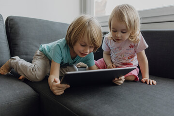 Brother and little sister using digital tablet on the couch at home
