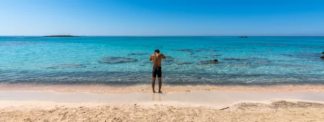 Foto auf Acrylglas Elafonissi Strand, Kreta, Griekenland Black boy by the sea. Spectacular panorama of Elafonissi Beach in Crete with Turquoise Water and the famous pink sand. The man is wearing a swimsuit and is sunny-side up. Banner.