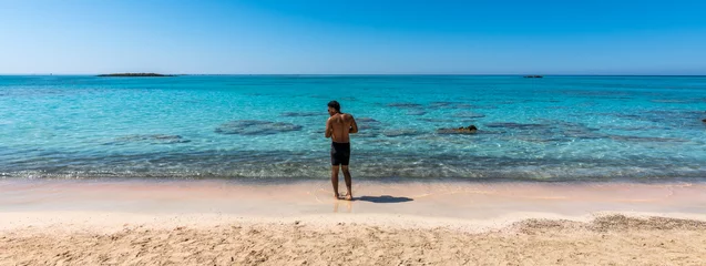 Photo sur Plexiglas  Plage d'Elafonissi, Crète, Grèce Black boy by the sea. Spectacular panorama of Elafonissi Beach in Crete with Turquoise Water and the famous pink sand. The man is wearing a swimsuit and is sunny-side up. Banner.