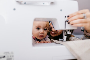 Little daughter watching her mother using sewing machine at home