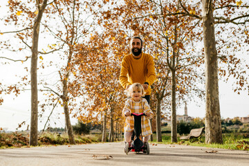 Father and daughter enjoying a morning day in the park in autumn, girl on scooter