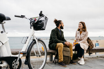 Couple sitting on a bench at beach promenade next to e-bike talking - obrazy, fototapety, plakaty
